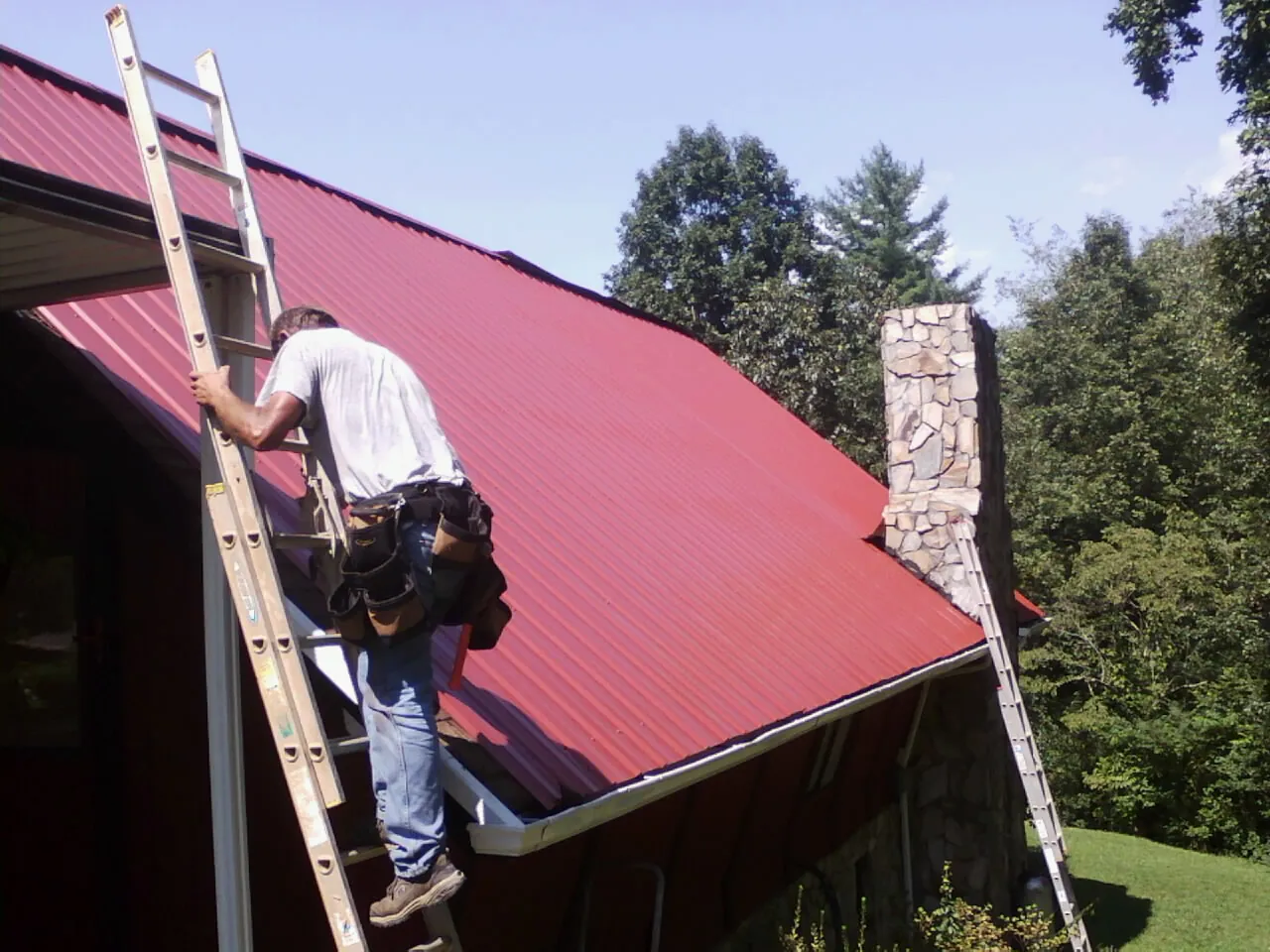 Roofer on a ladder beside a red metal roof and stone chimney — Soffit & Fascia Repair by Roofing Brothers in Mottville